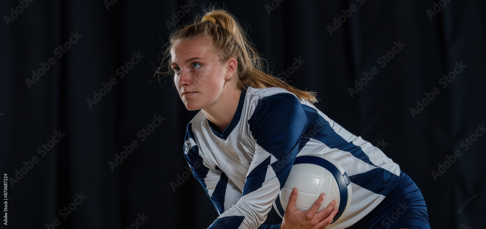 Obraz premium A young female volleyball player stands confidently with a volleyball in her hands. She is dressed in a team uniform and appears focused and ready for the game.