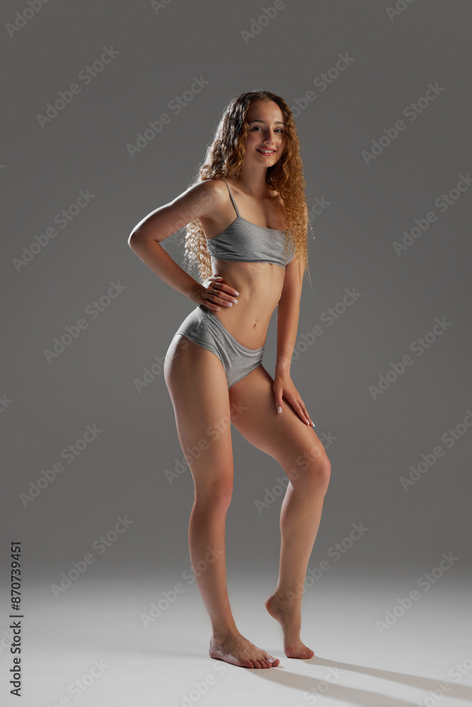 Fototapeta premium Full-length image of beautiful young girl showing her perfect fit body, model posing in underwear against grey studio background. Concept of female beauty, body and skin care, fitness, wellness