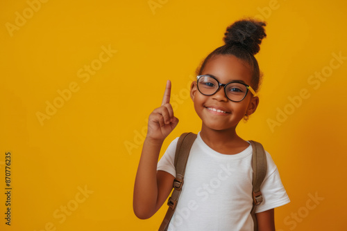 A happy smiley schoolgirl stands on a yellow empty background with a backpack pointing finger up at empty copyspace for text.