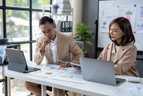 Two people are sitting at a desk with a white board behind them