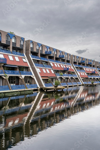 A picturesque scene showing a row of distinctive waterfront homes, their reflections perfectly mirrored in the calm waters of the canal under a cloudy sky in Rotterdam