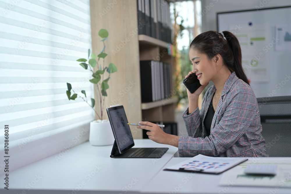 Young Professional Woman Working on Laptop and Talking on Phone in Modern Office Setting Young Professional Woman Working on Laptop and Talking on Phone in Modern Office Setting