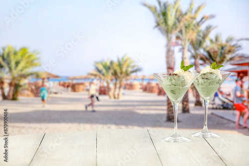 Fototapeta Naklejka Na Ścianę i Meble -  Mint and vanilla ice cream in glass with on wooden tabletop and blurred holiday resort background. Delicious sweet dessert on beach promenade. Tropical and exotic summer vacation 