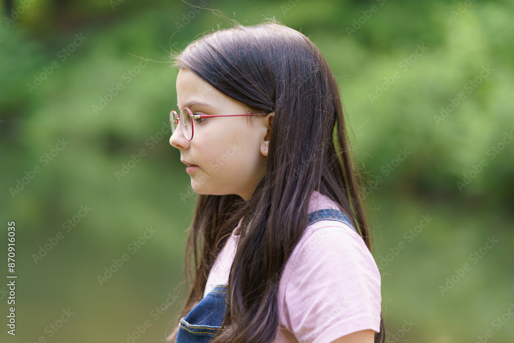 A little cute teenage girl in glasses stands near a pond. Child in denim overalls. Happy childhood concept A little cute teenage girl in glasses stands near a pond. Child in denim overalls. Happy childhood concept