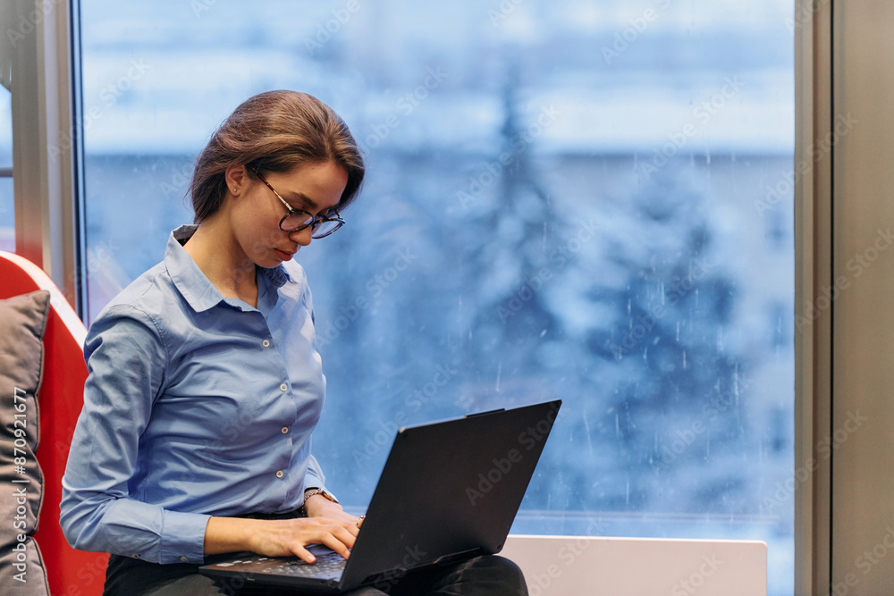 A businesswoman utilizes her laptop while seated by the window of a large corporate building, offering a picturesque view of the city skyline as her backdrop. A businesswoman utilizes her laptop while seated by the window of a large corporate building, offering a picturesque view of the city skyline as her backdrop.