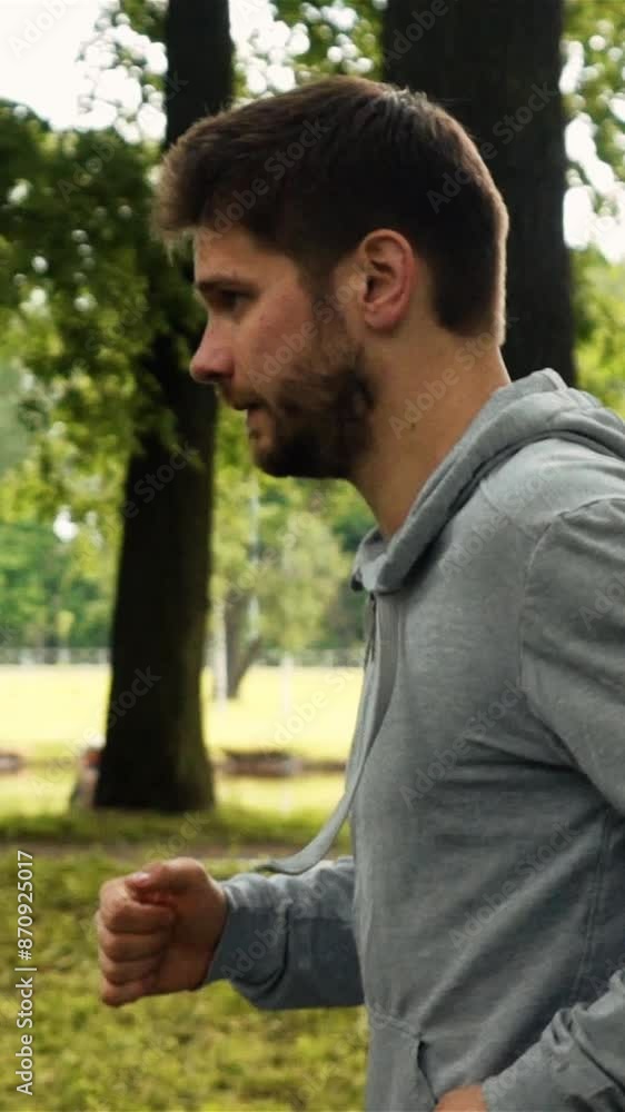 attractive powerful sportsmanman running in the park in gray jacket. Profile steadicam shot. Green trees background. Extreme slow motion footage. Vertical shot. attractive powerful sportsmanman running in the park in gray jacket. Profile steadicam shot. Green trees background. Extreme slow motion footage. Vertical shot.