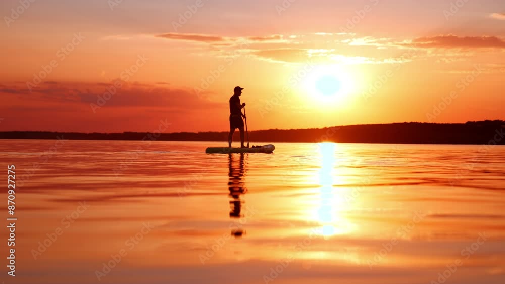 Lonely man going sup boarding on the river at sunset. Orange sky and waterscape with black silhouette of a man rowing on sup board. Lonely man going sup boarding on the river at sunset. Orange sky and waterscape with black silhouette of a man rowing on sup board.