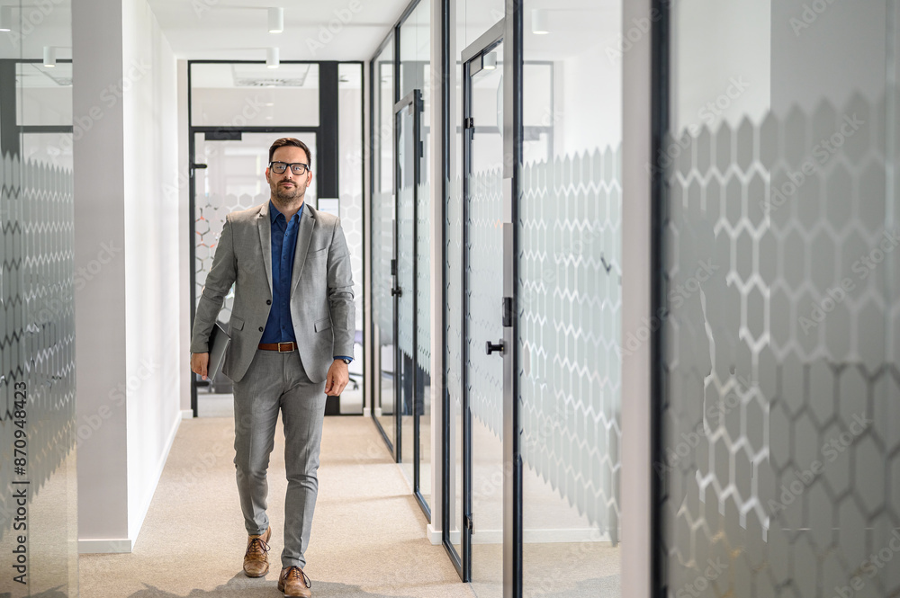Portrait of serious male manager in glasses holding laptop and walking confidently in office corridor Portrait of serious male manager in glasses holding laptop and walking confidently in office corridor