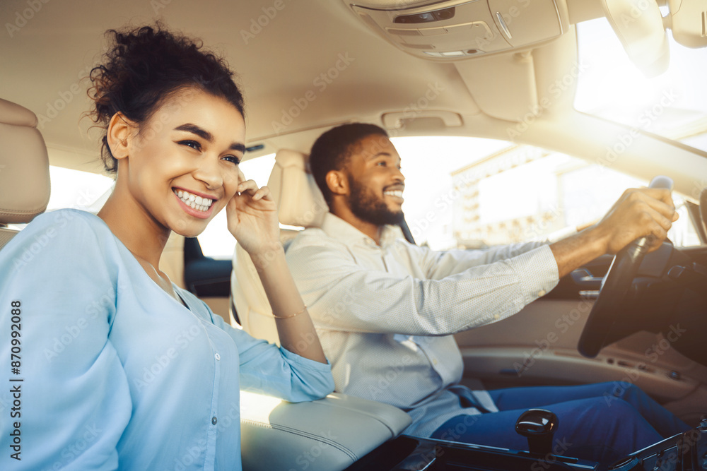 African American happy couple enjoys a car ride on a sunny day. The woman, with a big smile on her face, is in the passenger seat, while the man, wearing a white shirt, is driving. African American happy couple enjoys a car ride on a sunny day. The woman, with a big smile on her face, is in the passenger seat, while the man, wearing a white shirt, is driving.