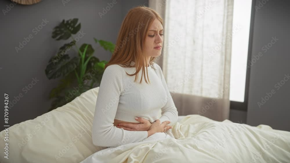 A pained young caucasian woman with red hair sits on a bed in an indoor bedroom setting, clutching her stomach. A pained young caucasian woman with red hair sits on a bed in an indoor bedroom setting, clutching her stomach.