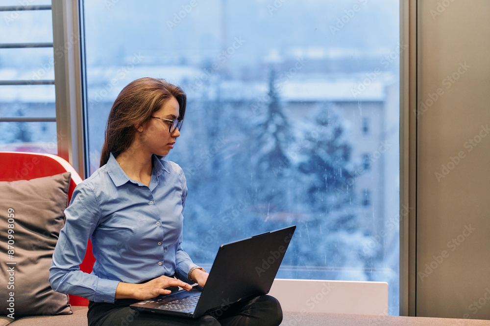 A businesswoman utilizes her laptop while seated by the window of a large corporate building, offering a picturesque view of the city skyline as her backdrop. A businesswoman utilizes her laptop while seated by the window of a large corporate building, offering a picturesque view of the city skyline as her backdrop.