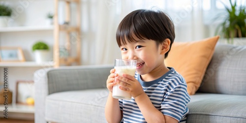 Cheerful young child in casual clothing sitting on couch in cozy living room, drinking fresh milk from blue cup.