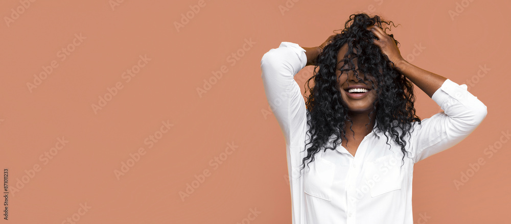 Smiling young Black woman with long, curly hair in front of a peach wall. She is wearing a white collared shirt and her arms are raised above her head as she runs her hands through her hair. Smiling young Black woman with long, curly hair in front of a peach wall. She is wearing a white collared shirt and her arms are raised above her head as she runs her hands through her hair.