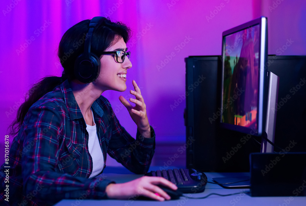 A woman with headphones on is playing a video game on a computer. The woman is smiling and has her hand on her chin as she looks intently at the screen. The room is dimly lit with purple lights. A woman with headphones on is playing a video game on a computer. The woman is smiling and has her hand on her chin as she looks intently at the screen. The room is dimly lit with purple lights.
