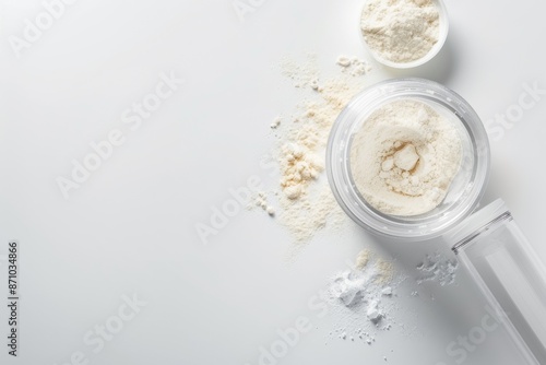 A high-angle photo of a whey protein powder container and a shaker bottle on a clean isolated white background. The powder is spilled out of the container and on the background