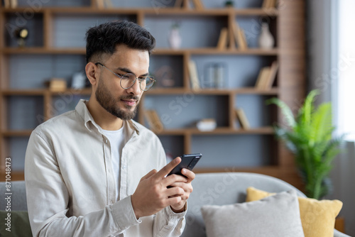 Close-up photo of a young Indian man sitting on the sofa at home in glasses and casual clothes, concentrating on using a mobile phone