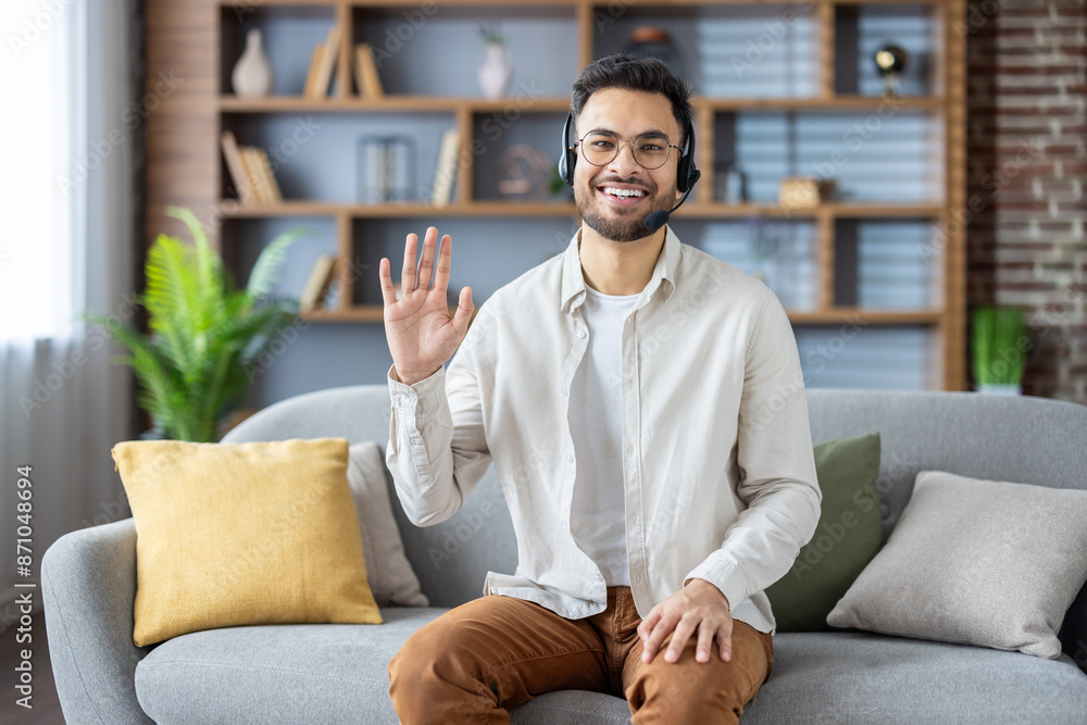 Portrait of a smiling young Indian man sitting on the couch at home wearing glasses and a headset, talking to the camera, waving and saying hello Portrait of a smiling young Indian man sitting on the couch at home wearing glasses and a headset, talking to the camera, waving and saying hello