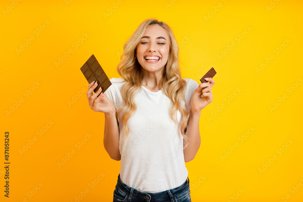 This image features a young woman with long blonde hair smiling widely while holding two chocolate bars in her hands. She is standing in front of a bright yellow background. This image features a young woman with long blonde hair smiling widely while holding two chocolate bars in her hands. She is standing in front of a bright yellow background.