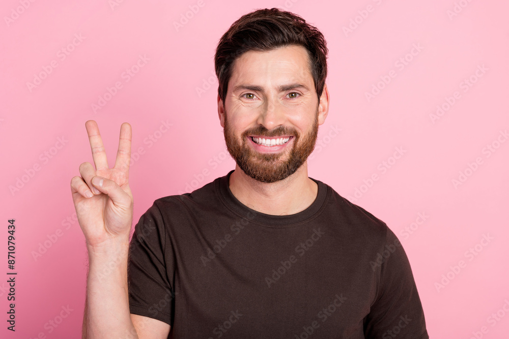 Photo of good mood optimistic guy with stubble brunet hair dressed brown t-shirt showing v-sign symbol isolated on pink color background Photo of good mood optimistic guy with stubble brunet hair dressed brown t-shirt showing v-sign symbol isolated on pink color background