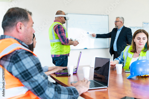 A group of construction workers are sitting around a table having a meeting about virtual reality. They are wearing , hard hats and safety vests. There are three men and two women in the group.
