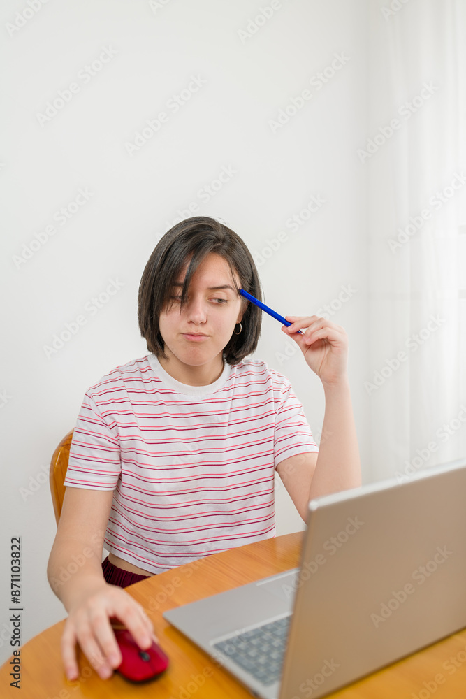 University female student deep in thought while studying at home. The student is sitting at a wooden table with an open laptop, suggesting a focus on online learning background white wall.