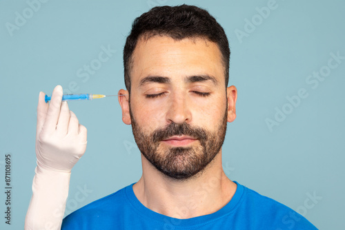 Handsome bearded middle-aged man with closed eyes and unrecognizable doctor or nurse hand with syringe over blue studio background. Male, men's cosmetology, aesthetic medicine concept