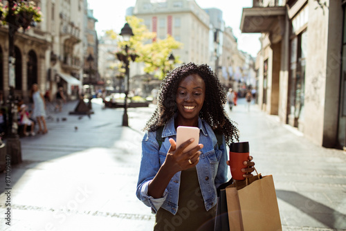 Happy Black woman using smartphone while holding coffee and shopping bags on urban street