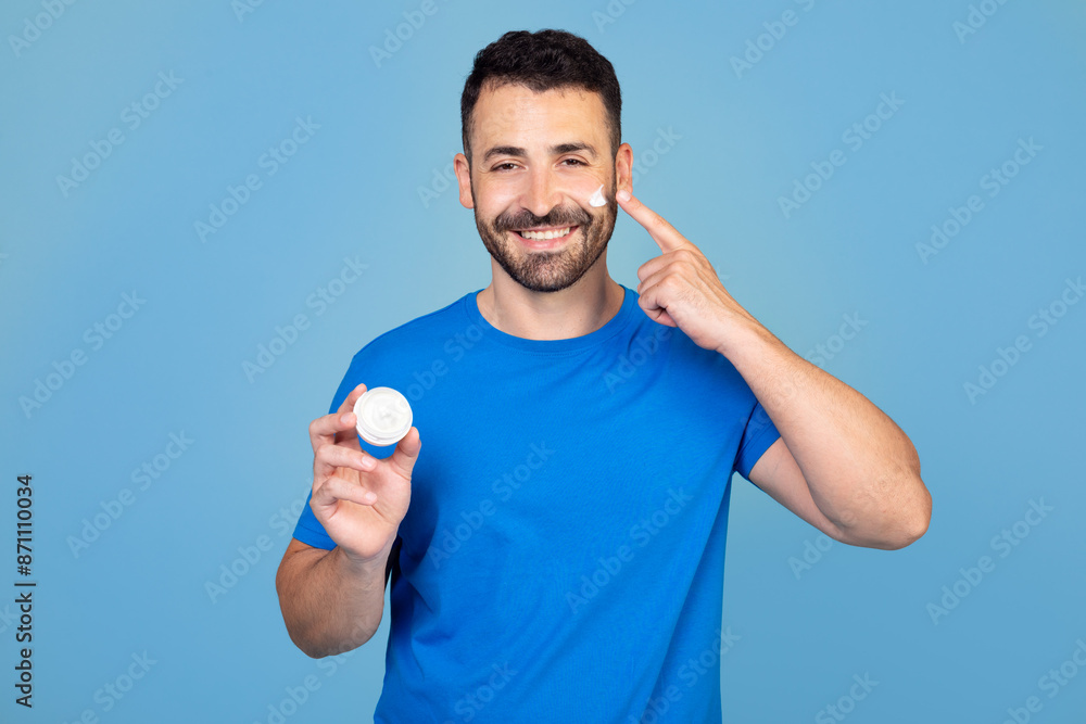 Youthful man smiles with joy as he introduces a beauty cream to his skincare routine. Happy young man pampering his face with a moisturizing product to achieve a glowing and radiant skin tone. Youthful man smiles with joy as he introduces a beauty cream to his skincare routine. Happy young man pampering his face with a moisturizing product to achieve a glowing and radiant skin tone.