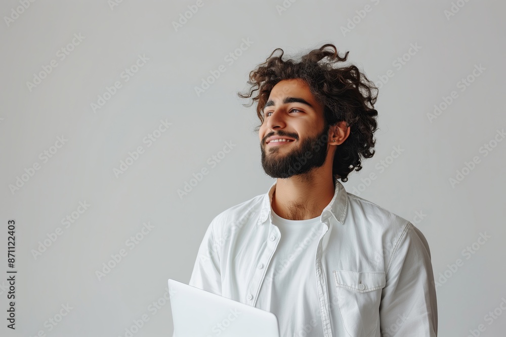 Obraz premium A man with a beard and a white shirt is smiling and holding a laptop. Concept of happiness and contentment, as the man is enjoying his time with the laptop