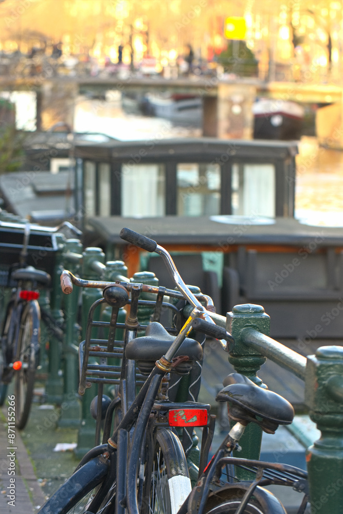 Cycles, bicycles and bikes parked on the road and against the wall on the bridge over the canal in Amsterdam city. Picturesque sunny town landscape in the Netherlands with a view of river Amstel. Cycles, bicycles and bikes parked on the road and against the wall on the bridge over the canal in Amsterdam city. Picturesque sunny town landscape in the Netherlands with a view of river Amstel.