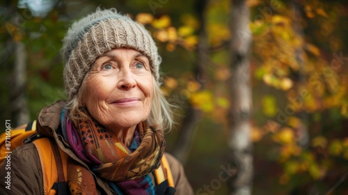 An elderly woman with gray hair, wearing a knit beanie and scarf, stands in an autumn forest. Her face emanates a serene and reflective expression, with colorful foliage behind.