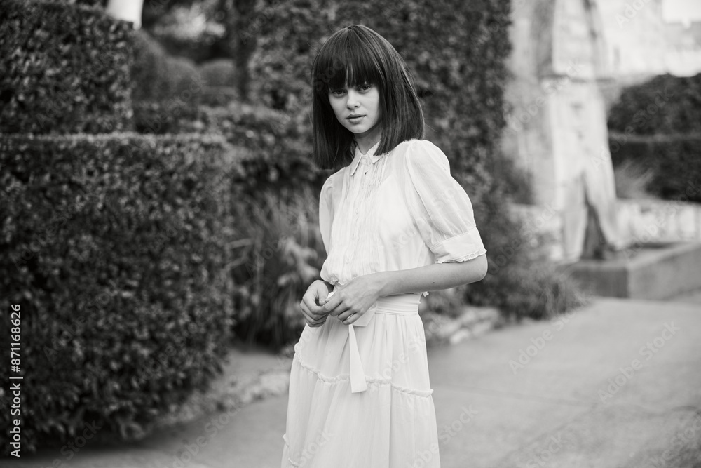 Elegant woman in white dress standing gracefully against lush garden backdrop Elegant woman in white dress standing gracefully against lush garden backdrop