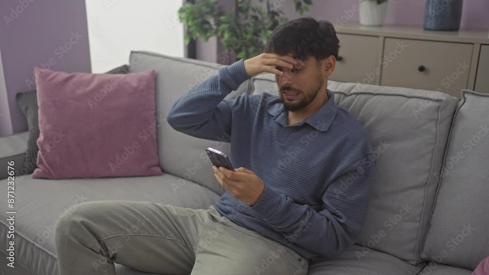 Stressed young arab man sitting on sofa at home, expressing frustration and surprise while using smartphone
