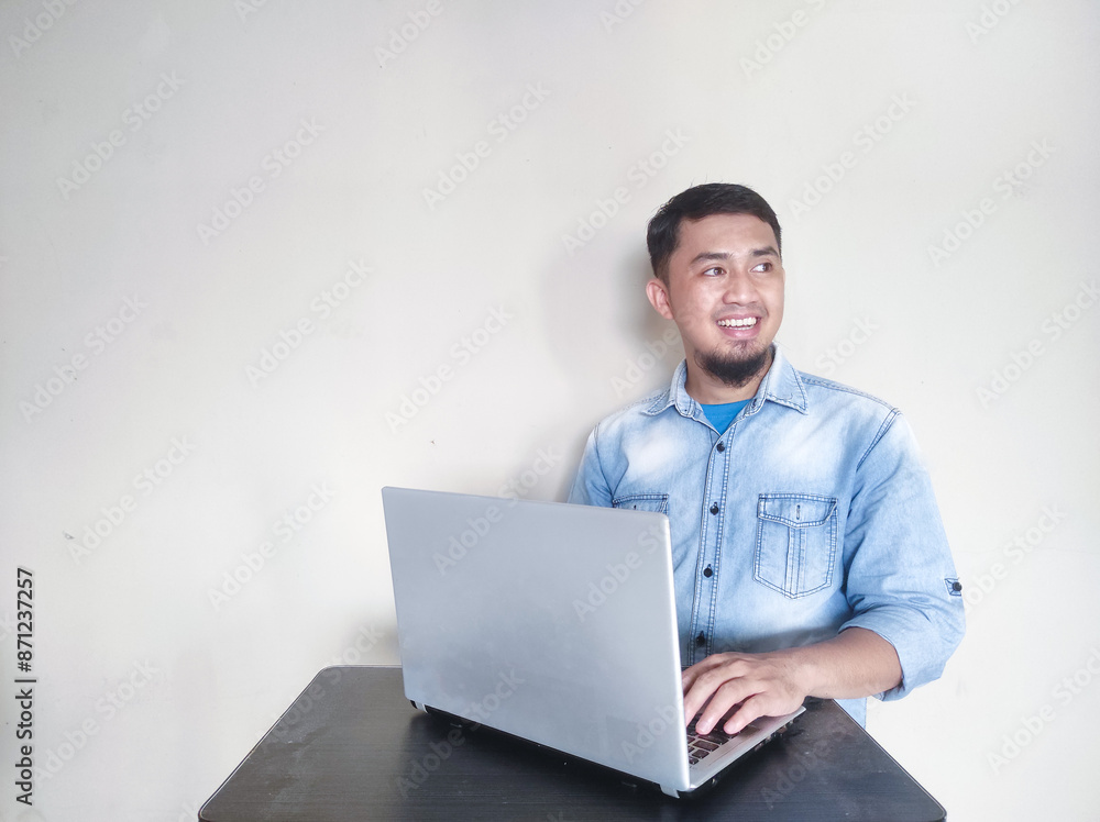 Young Asian man feeling happy and smiling while working on laptop, while looking at empty area beside him Young Asian man feeling happy and smiling while working on laptop, while looking at empty area beside him