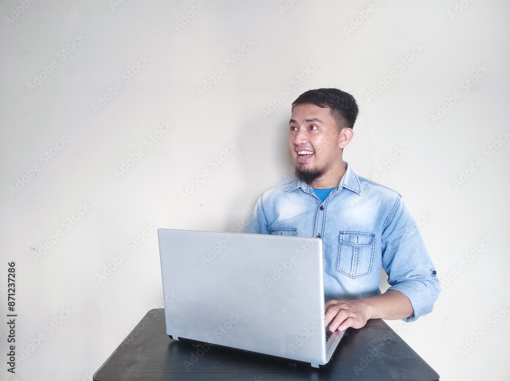 Young Asian man feeling happy and smiling while working on laptop, while looking at empty area beside him Young Asian man feeling happy and smiling while working on laptop, while looking at empty area beside him