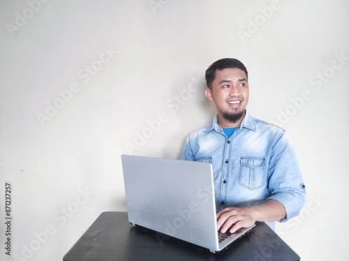 Young Asian man feeling happy and smiling while working on laptop, while looking at empty area beside him