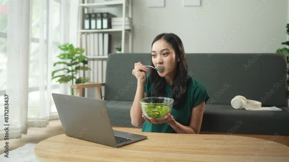 Young professional enjoys a healthy green salad while working remotely on a laptop from home. Seated at a table in a cozy living room with natural light and minimalist decor Young professional enjoys a healthy green salad while working remotely on a laptop from home. Seated at a table in a cozy living room with natural light and minimalist decor