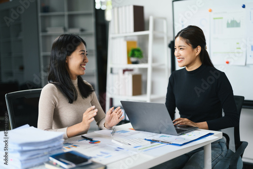 Two Businesswomen Collaborating in Modern Office with Laptops and Documents, Smiling and Engaged in Productive Discussion