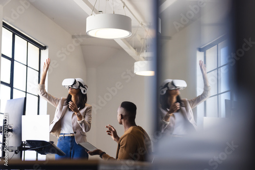 Using VR headset in office, woman exploring virtual environment while colleague observes photo