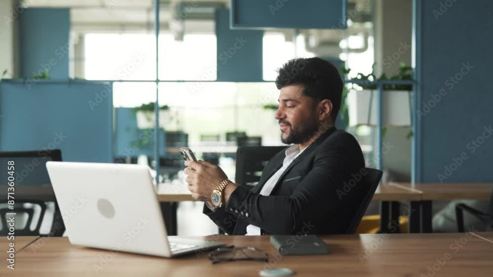 portrait of a handsome, stylish young man of Arab descent sitting in a modern business center office with a laptop, texting on his smartphone in social media and messengers portrait of a handsome, stylish young man of Arab descent sitting in a modern business center office with a laptop, texting on his smartphone in social media and messengers