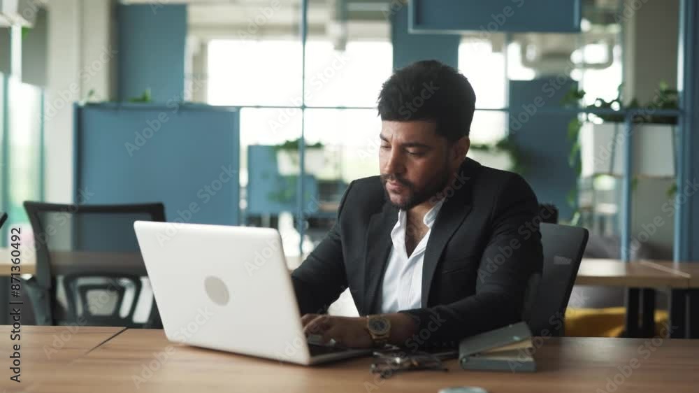 portrait of a handsome, stylish young man of Arab descent sitting in a modern office of a business center, working intently on his laptop and contemplating portrait of a handsome, stylish young man of Arab descent sitting in a modern office of a business center, working intently on his laptop and contemplating