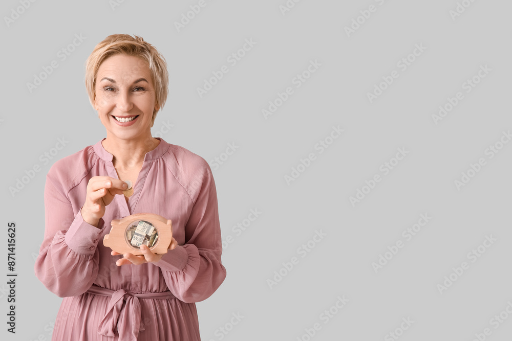 Beautiful mature woman putting coins into wooden piggy bank on grey background Beautiful mature woman putting coins into wooden piggy bank on grey background
