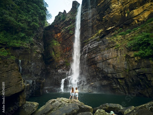 A couple of young women stand against a tall tropical waterfall and hold hands enjoying the view photo