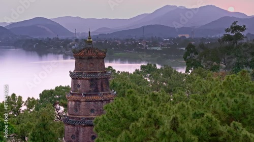 This footage captures a peaceful sunset over Hue, Vietnam, featuring a beautiful pagoda surrounded by lush trees, with the city and distant mountains in the background. 