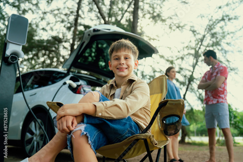 Little boy portrait sitting on camping chair with his family in background. Road trip travel with alternative energy charging station for eco-friendly car concept. Perpetual © Summit Art Creations