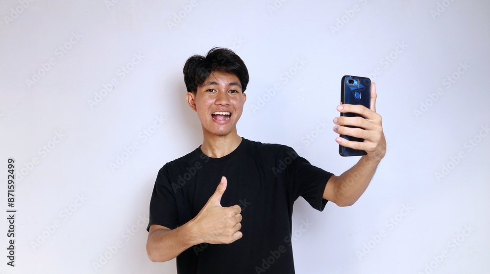 Happy young Asian man wearing black shirt gesturing okay while holding smartphone Happy young Asian man wearing black shirt gesturing okay while holding smartphone