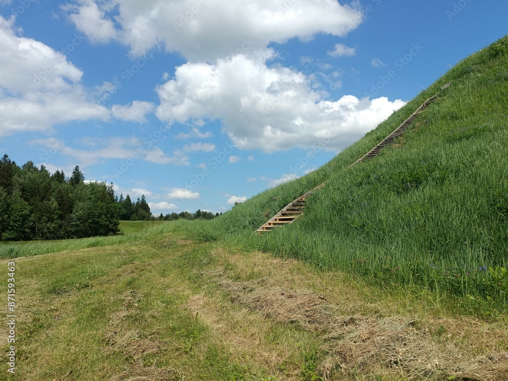 Bubiai hill during sunny day. Small hill. Grass is growing on hill. Staircase leading to the top. Sunny day with white and gray clouds in sky. Nature. Bubiu piliakalnis. Bubiai hill during sunny day. Small hill. Grass is growing on hill. Staircase leading to the top. Sunny day with white and gray clouds in sky. Nature. Bubiu piliakalnis.