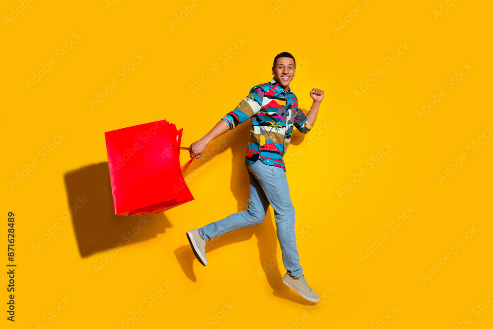 Full length photo of nice young male running shopping bags dressed stylish colorful garment isolated on yellow color background Full length photo of nice young male running shopping bags dressed stylish colorful garment isolated on yellow color background