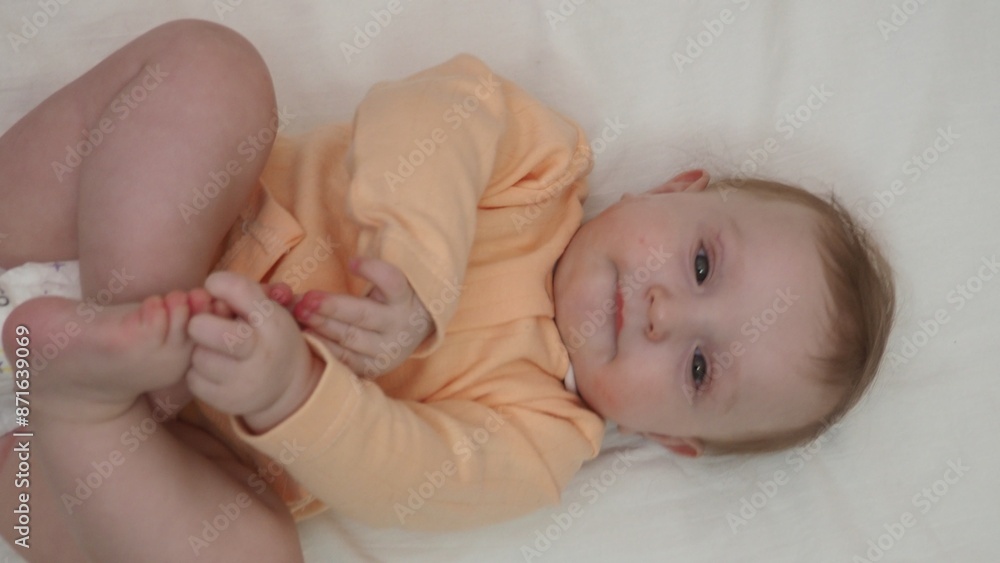 Close up sweet beautiful newborn baby in bodysuit and dry diaper lying down on white bedsheets, laughing, looks carefree plays, touches with toes. Babyhood, healthcare, paediatrics, childcare concept Close up sweet beautiful newborn baby in bodysuit and dry diaper lying down on white bedsheets, laughing, looks carefree plays, touches with toes. Babyhood, healthcare, paediatrics, childcare concept