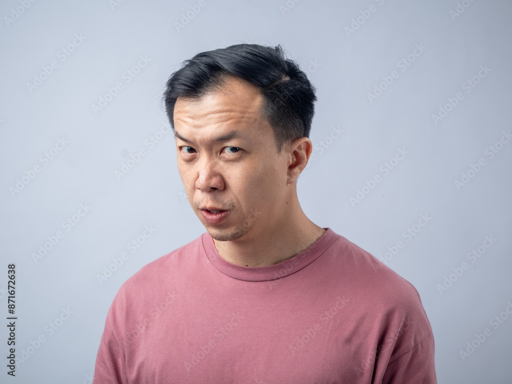 An Asian man in a pink shirt makes a skeptical and uncomfortable expression, as if reacting to something unpleasant. The plain light blue background highlights his displeased